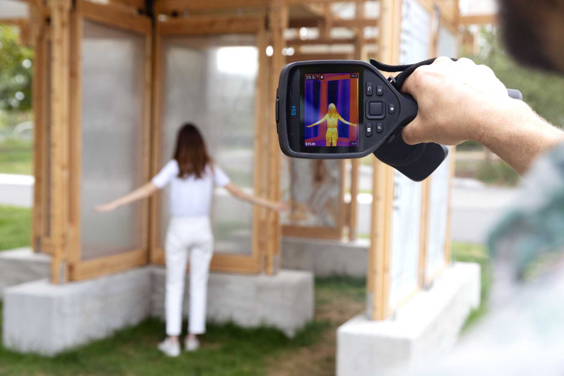 Person with arms outstretched being scanned by a FLIR thermal camera showing a heat map with temperature readings from 20°C to 31°C.